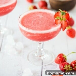 A refreshing glass of strawberry Frosé, elegantly garnished with a strawberry slice, surrounded by fresh strawberries and glistening ice cubes on a pristine white surface, highlighting its cool appeal.