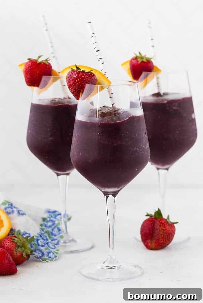 Refreshing Sangria Slushie 2 Three long stem wine glasses filled with vibrant red frozen sangria, garnished with fresh strawberries and orange slices. Paper straws are placed in each glass, ready for sipping on a warm day.