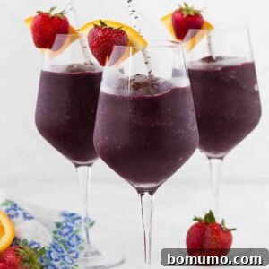 Three long stem wine glasses filled with frozen sangria, and topped with a fresh strawberry and orange slice. A paper straw is in each glass.