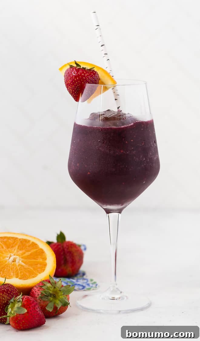 Refreshing Sangria Slushie 4 A long-stemmed wine glass brimming with icy red wine sangria slushie, exquisitely garnished with a fresh strawberry and a vibrant orange slice. A festive paper straw is nestled within the drink, ready to be enjoyed.