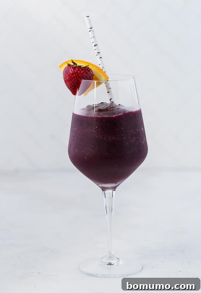 Refreshing Sangria Slushie 5 A long-stemmed wine glass filled with a deep red frozen sangria slushie, beautifully garnished with a fresh strawberry and an orange slice. A paper straw is ready for sipping.