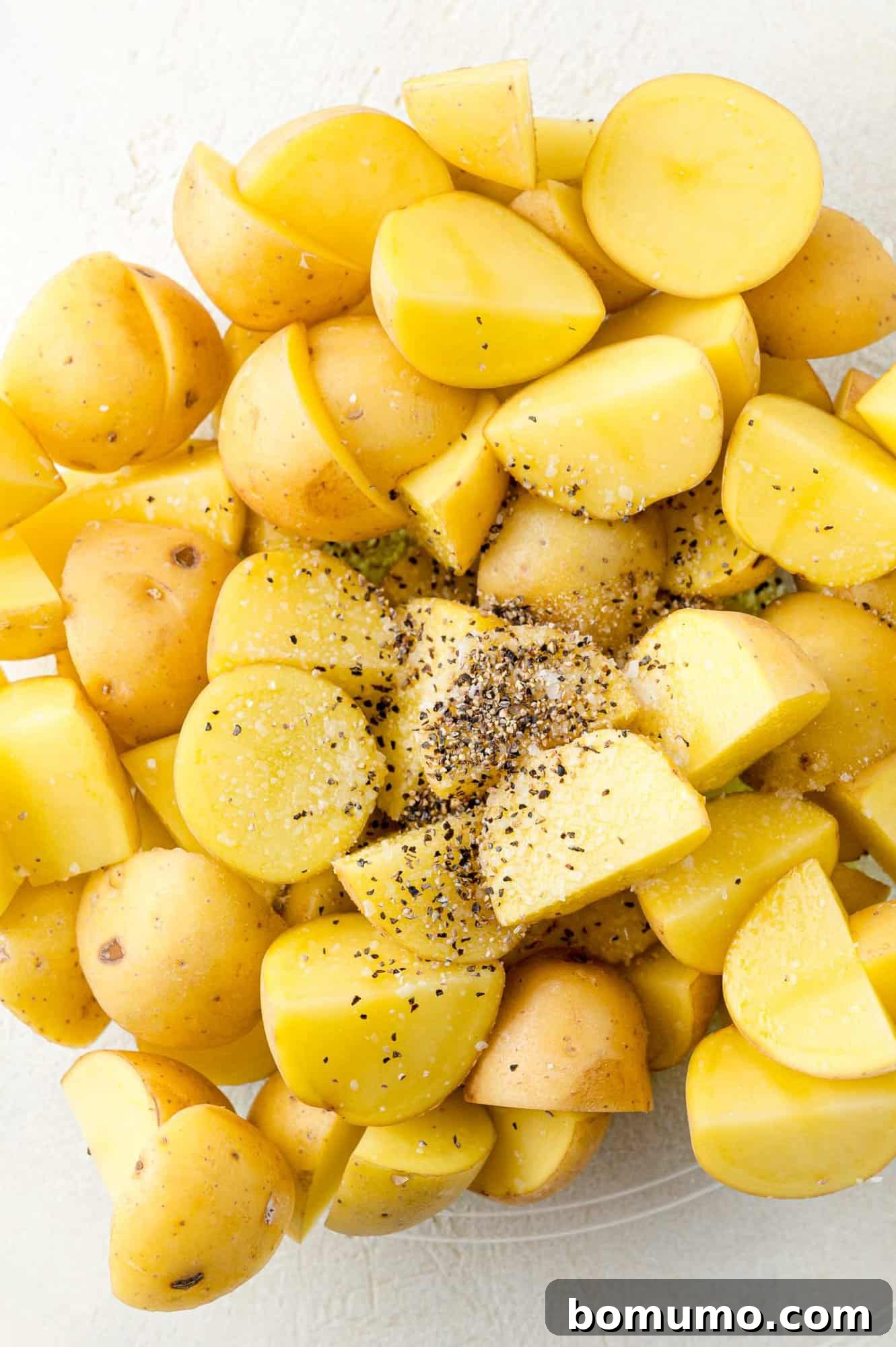 Freshly cut potatoes in a bowl, with olive oil, kosher salt, and black pepper being added, showcasing the seasoning process.