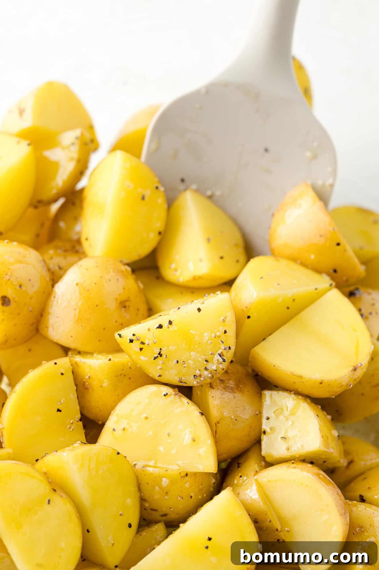 Halved baby potatoes are shown in a clear glass bowl, being thoroughly mixed with olive oil, salt, and pepper to ensure even coating.