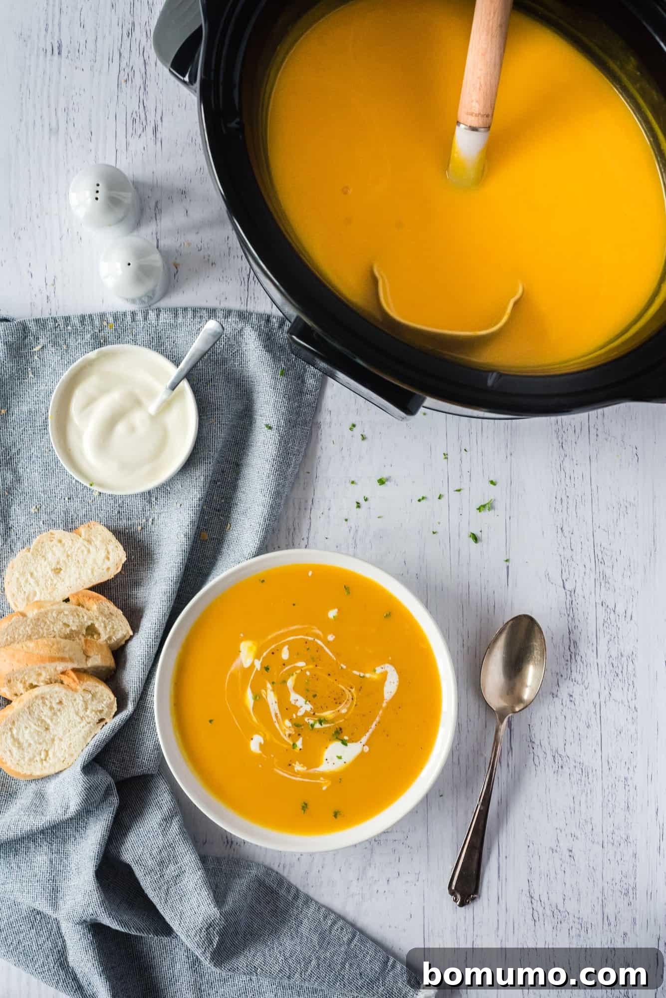 Overhead view of a bowl of pumpkin soup on a countertop next to a crockpot and slices of bread.