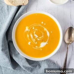 Overhead view of a bowl of pumpkin soup swirled with cream, next to bread slices and a spoon.