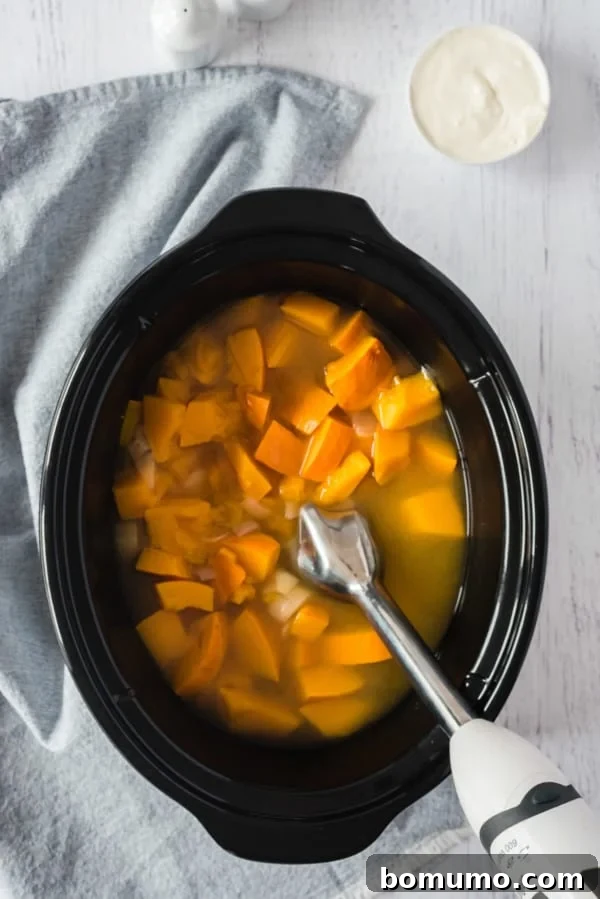 An immersion blender resting in a crockpot of cooked pumpkin soup ingredients.
