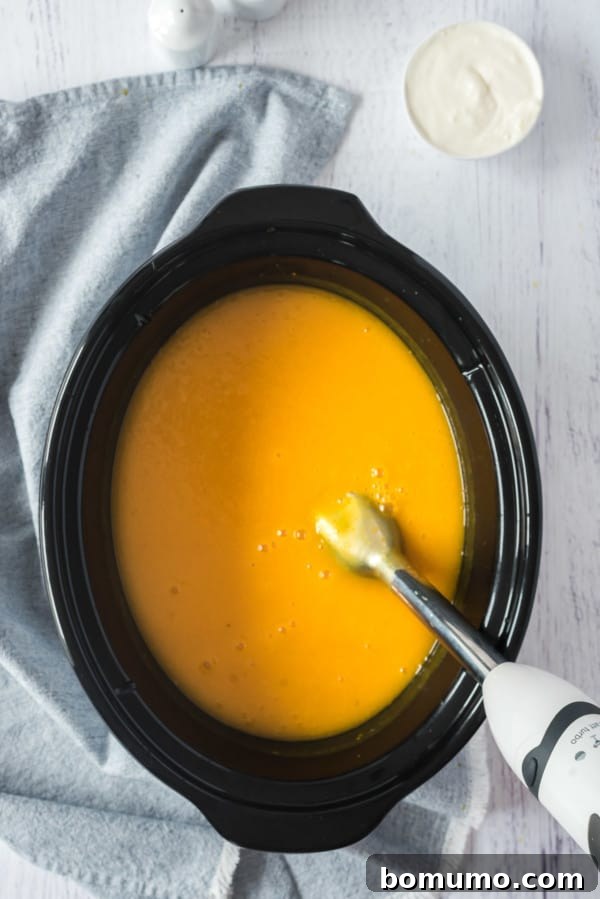 An immersion blender resting in a crockpot of pureed pumpkin soup.