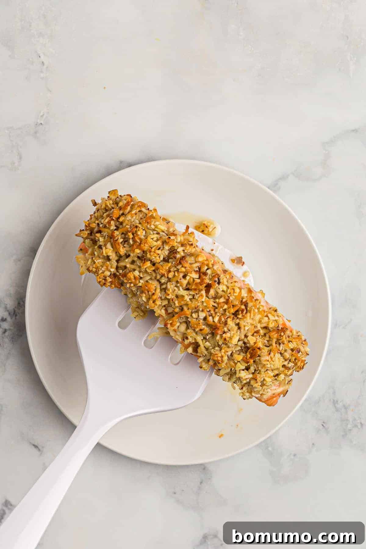 A perfectly cooked, flaky almond and Parmesan crusted salmon fillet resting on a spatula over a white marble background, with a white plate waiting underneath. The crust is golden and inviting.