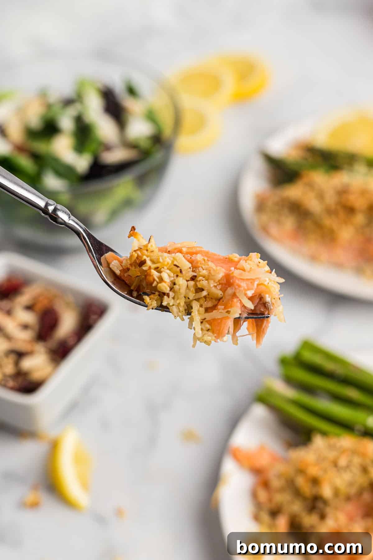 A close-up of a fork lifting a flaky piece of almond and Parmesan crusted salmon, with a complete plated meal and fresh asparagus visible in the softly blurred background.