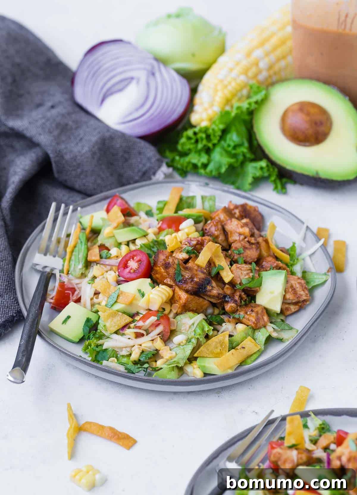 Colorful chopped salad on a grey plate. Salad includes bbq grilled chicken, lettuce, onion, tomato, crispy tortilla strips, avocado, corn and more! Whole ingredients are featured in the background of the image.