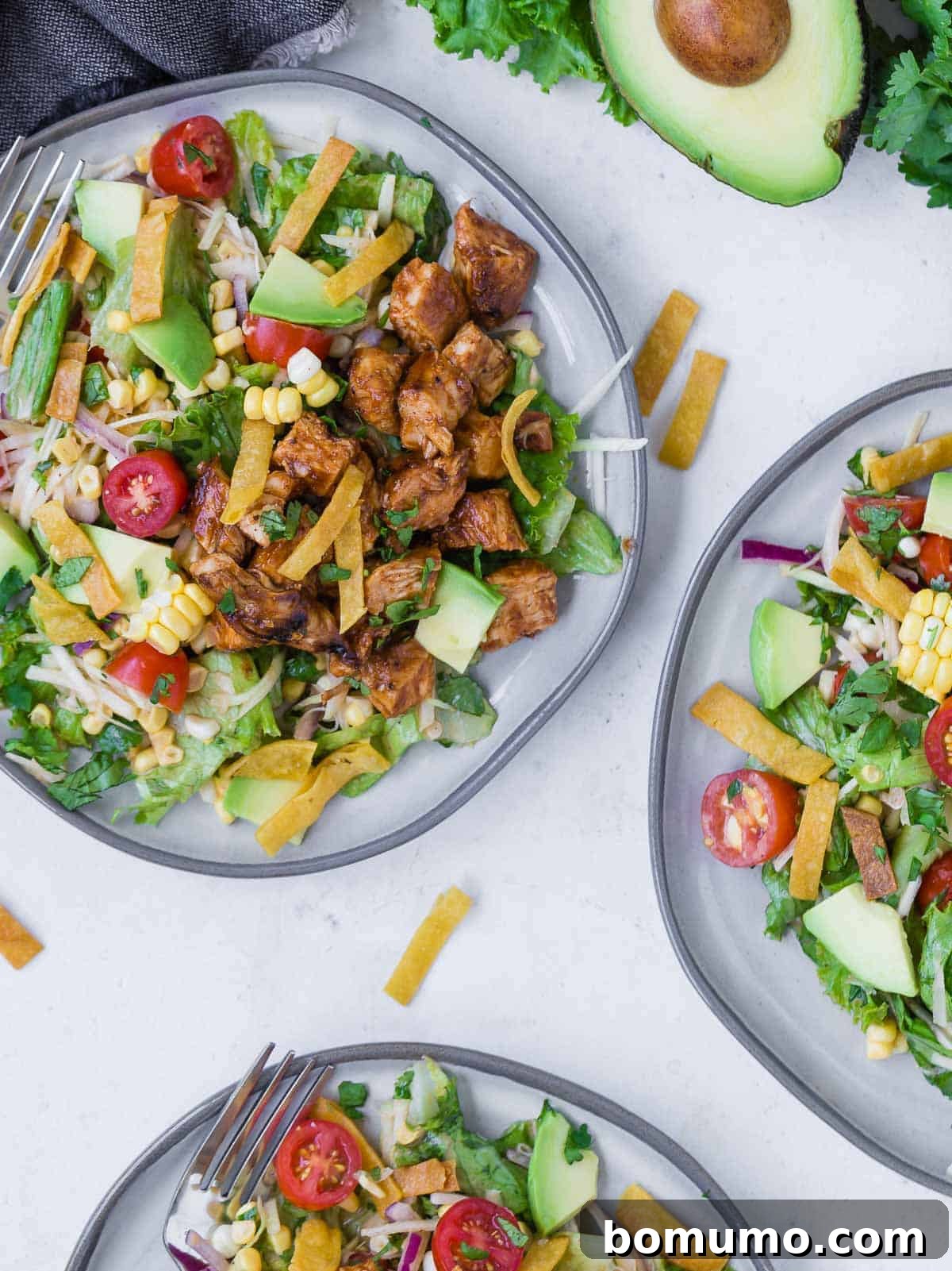 Overhead view of three plates of a colorful chopped salad on a grey plate. Salad includes bbq grilled chicken, lettuce, onion, tomato, crispy tortilla strips, avocado, corn and more!