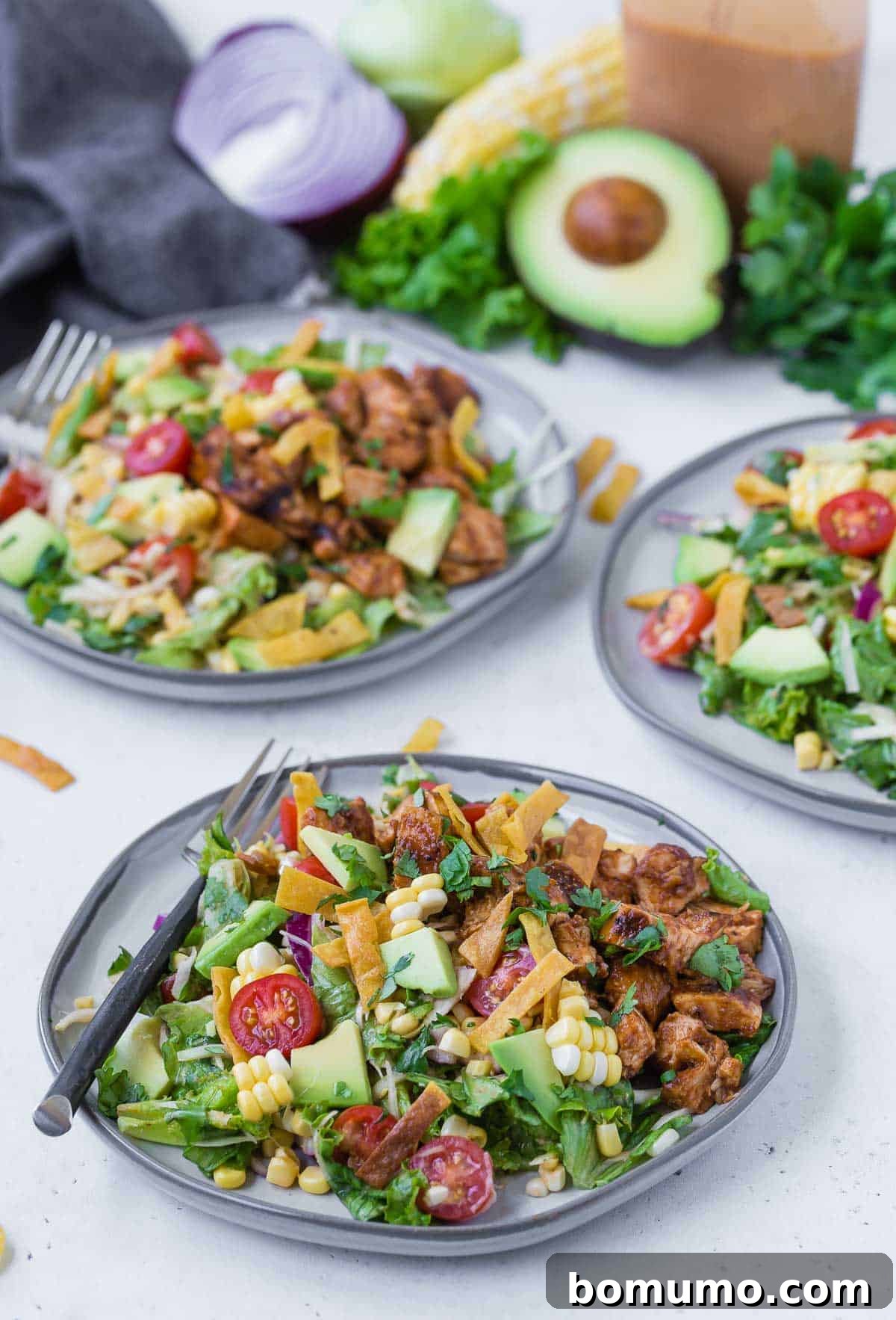 Three plates of a colorful chopped salad on a grey plates. Salad includes bbq grilled chicken, lettuce, onion, tomato, crispy tortilla strips, avocado, corn and more!