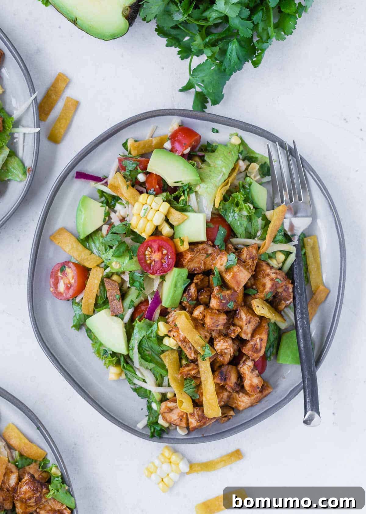 Overhead view of three plates of a colorful chopped salad on a grey plate. Salad includes bbq grilled chicken, lettuce, onion, tomato, crispy tortilla strips, avocado, corn and more!