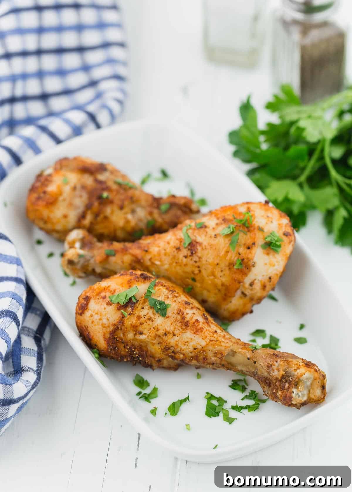 Three perfectly cooked chicken drumsticks arranged on a white serving platter against a white wooden background.