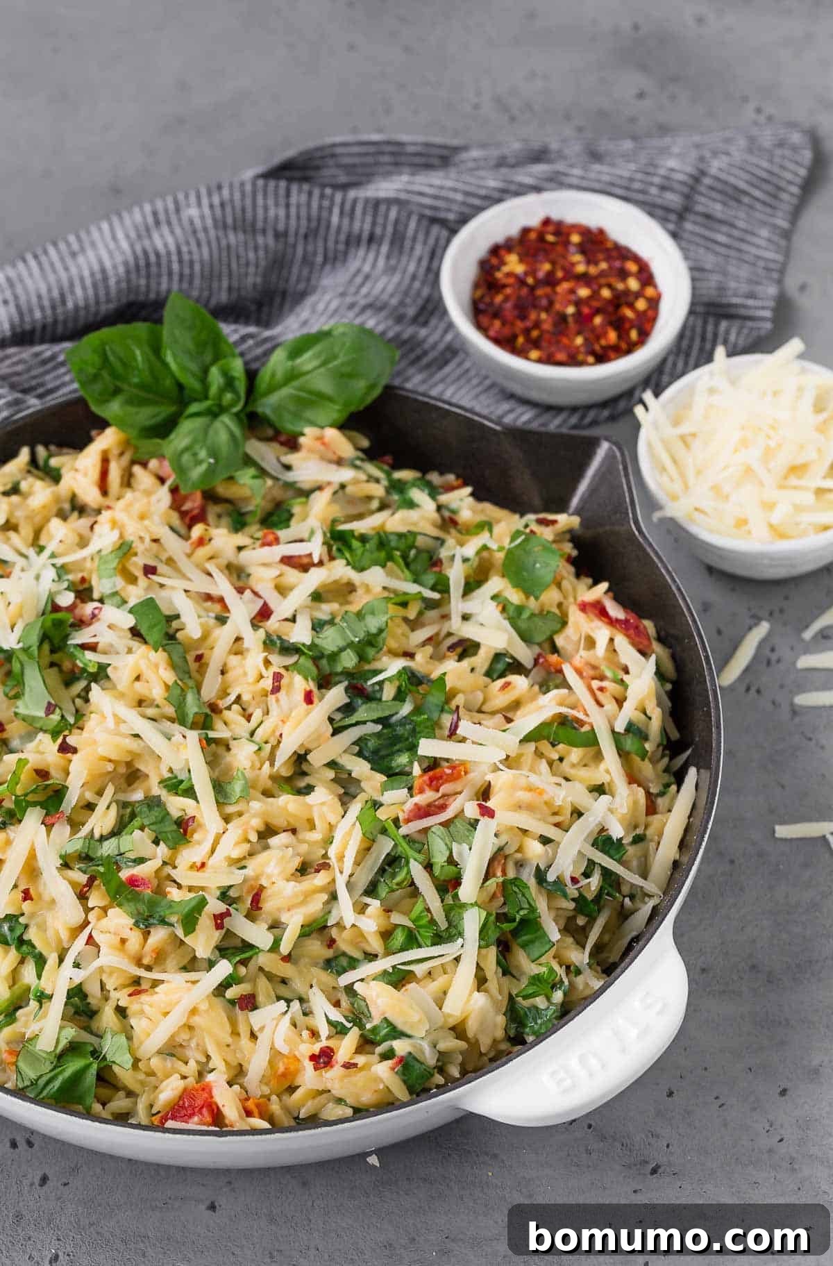 A striking white and black skillet rests on a textured grey surface, showcasing a generous portion of creamy orzo. The pasta is beautifully intertwined with fresh spinach, vibrant sun-dried tomatoes, and fragrant basil leaves.