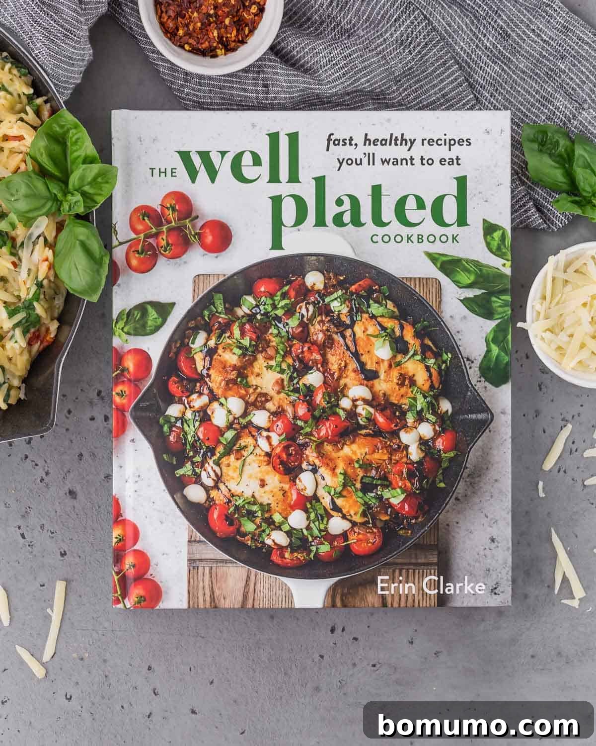 A copy of The Well Plated Cookbook rests on a simple gray surface. Surrounding it are a serving of creamy orzo pasta, a small bowl of shredded parmesan cheese, a small bowl of red pepper flakes, and vibrant fresh basil sprigs, hinting at the delightful flavors within the cookbook.