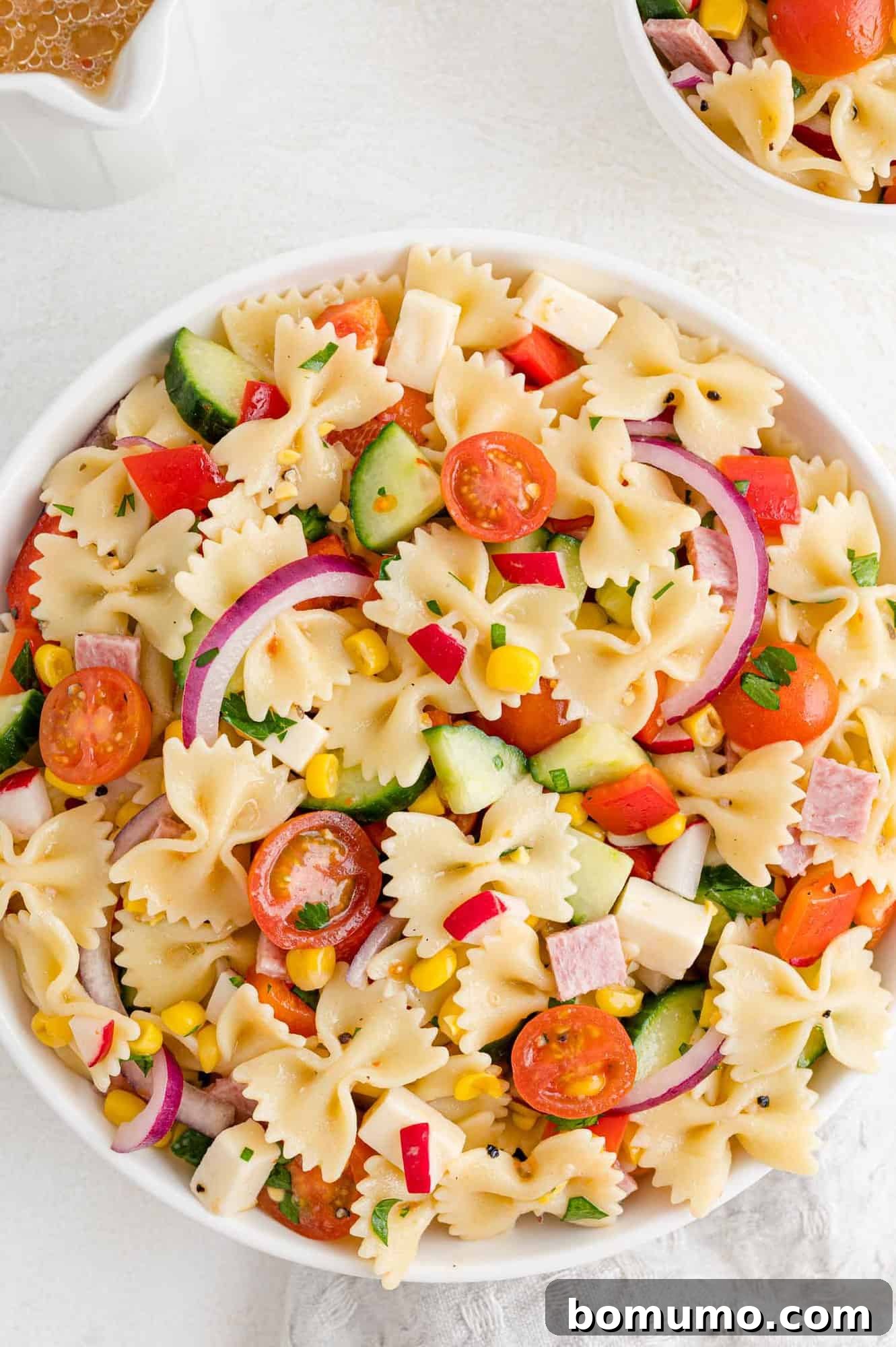 A refreshing summer pasta salad, featuring colorful bowtie pasta, crisp vegetables, and savory salami, beautifully arranged in a white round bowl, ready to be served.