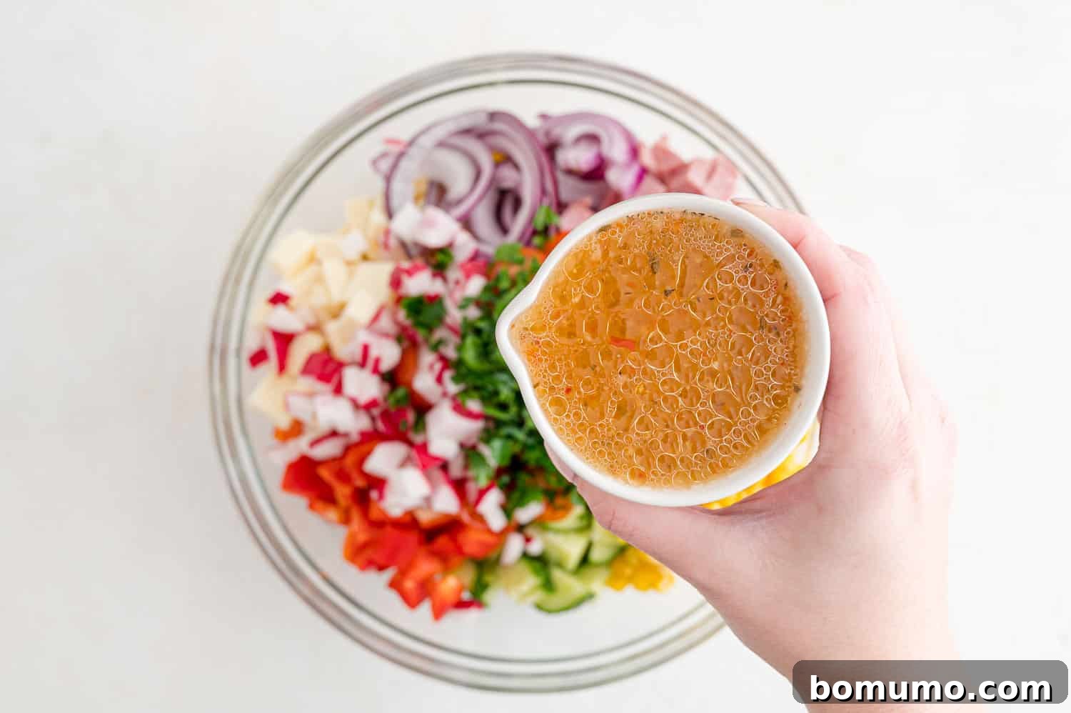 Italian dressing being generously drizzled over the prepared pasta and vegetables in a mixing bowl.