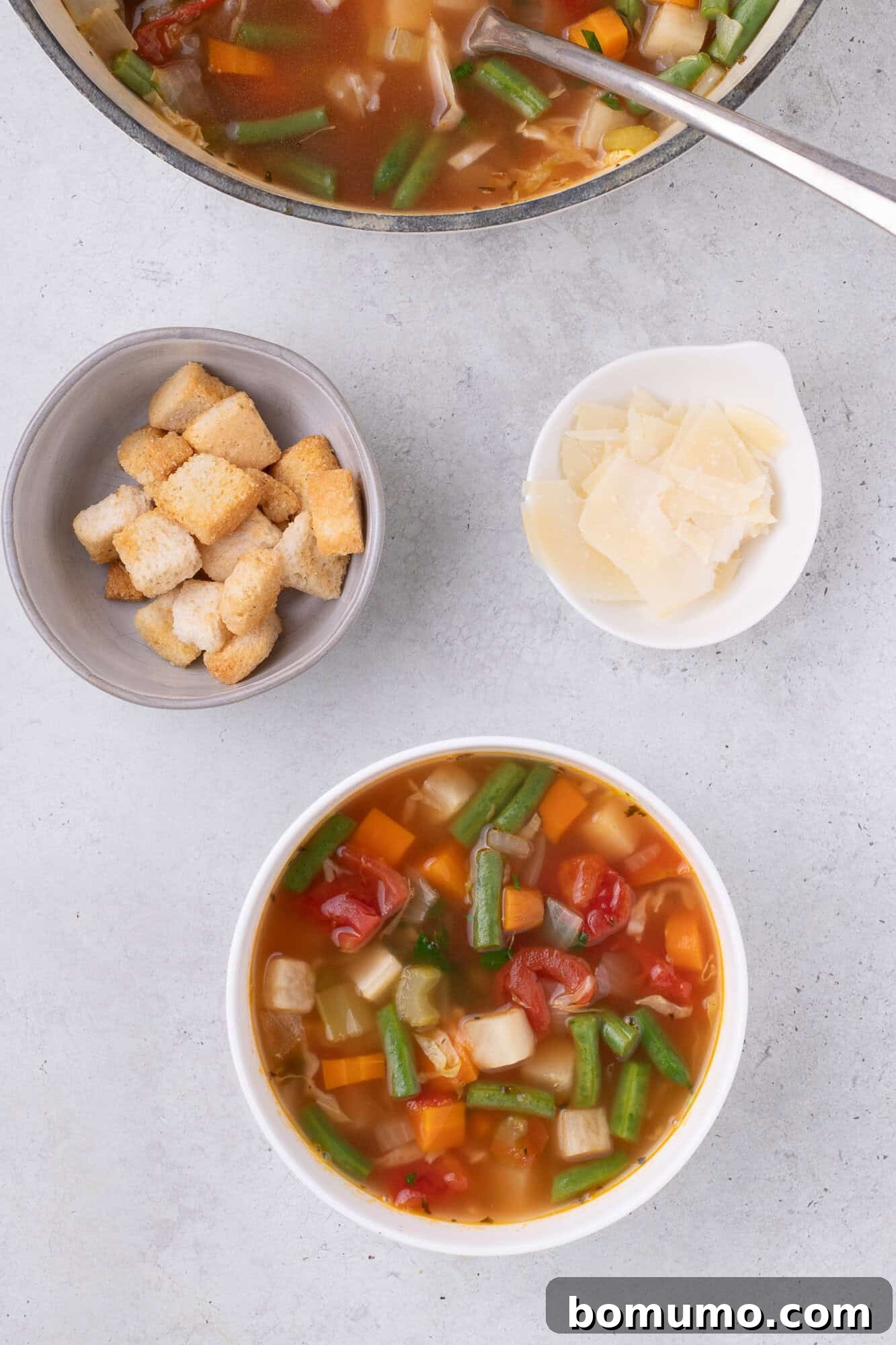 A small white bowl of vegetable soup, garnished with Parmesan cheese and a side of croutons.