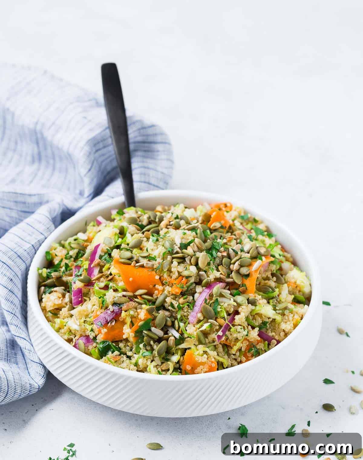 A white bowl filled with a colorful and hearty vegan quinoa salad, featuring prominent shredded Brussels sprouts, carrot ribbons, and red onions.