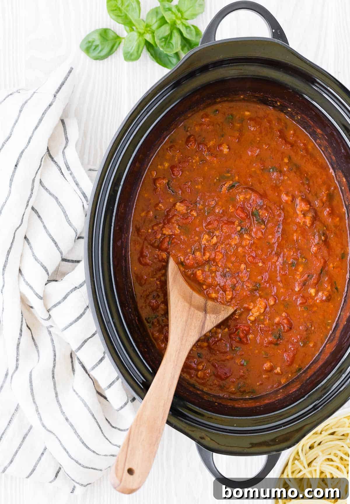 Rich spaghetti sauce simmering in a black slow cooker, with a wooden spoon resting inside, ready to be served over pasta.