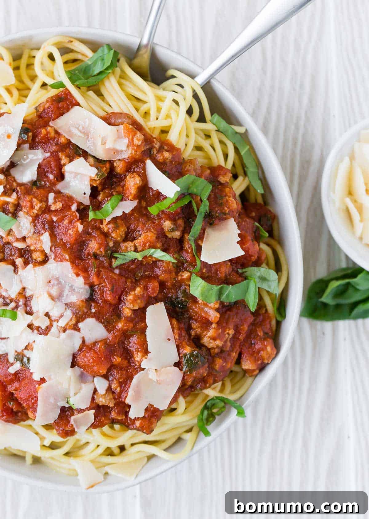 An inviting overhead shot of a large white bowl generously filled with perfectly cooked spaghetti, topped with savory meat sauce and fresh basil.