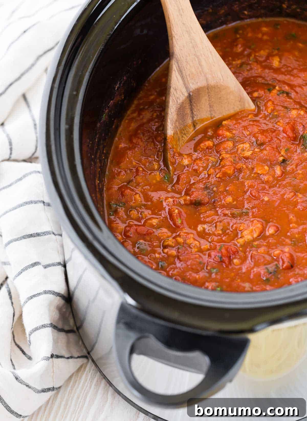 A wooden spoon rests inside a black slow cooker, stirring the rich, red spaghetti sauce, highlighting its thick texture.