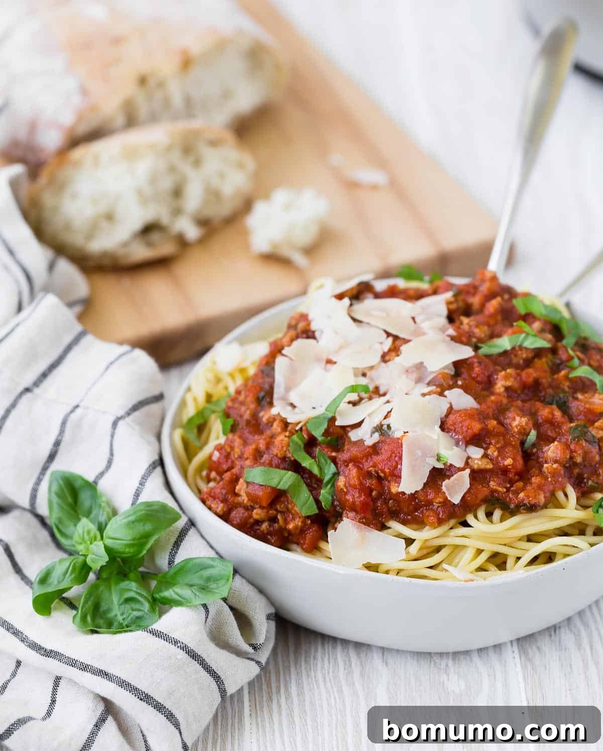 A beautifully plated bowl of spaghetti topped with a generous serving of red sauce, garnished with flakes of Parmesan cheese and fresh basil, with warm bread in the background.