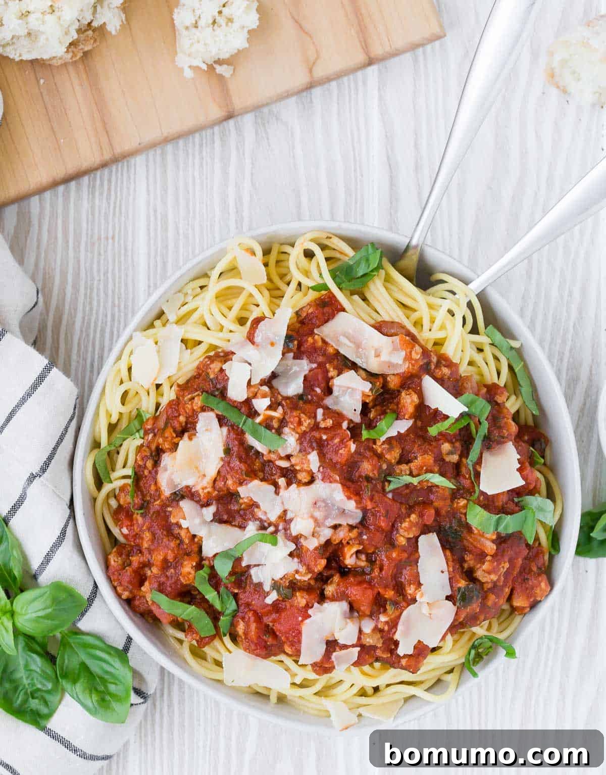 An inviting overhead view of a white bowl overflowing with spaghetti, generously topped with savory sauce and fresh Parmesan, with elegant silver serving utensils.