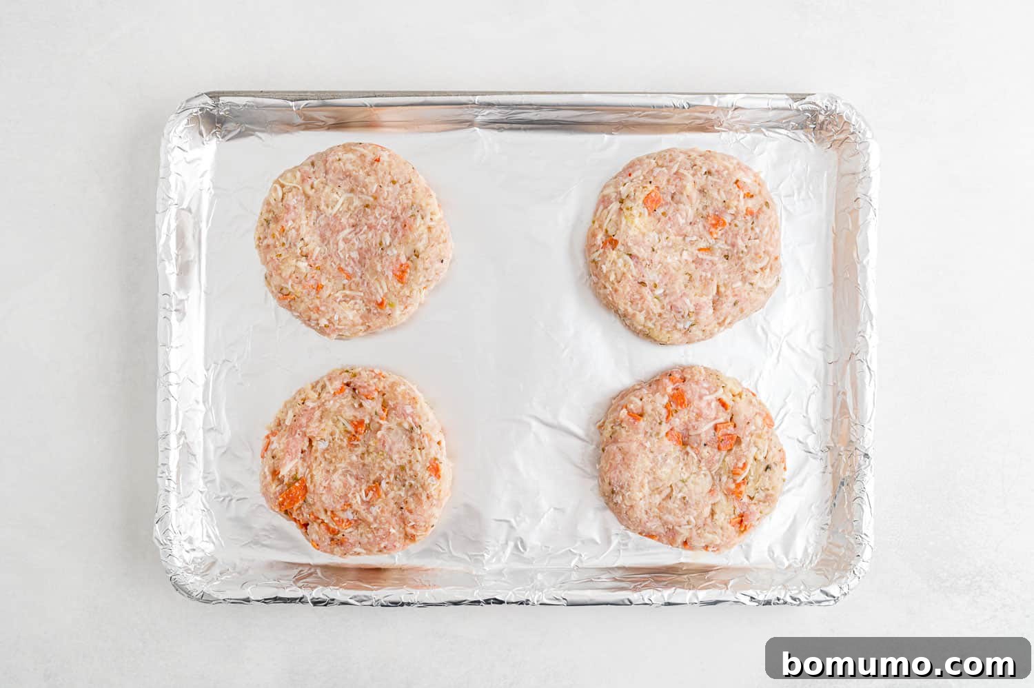 Four perfectly shaped, uncooked turkey burger patties resting on a baking sheet, ready for broiling.