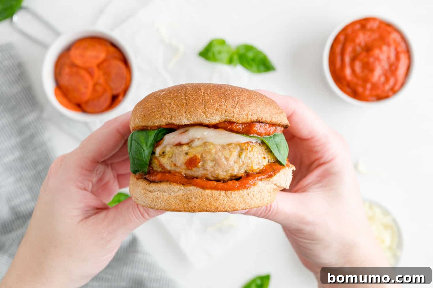 A delicious pizza turkey burger, perfectly assembled and held in two hands, showcasing its inviting layers and fresh basil topping.