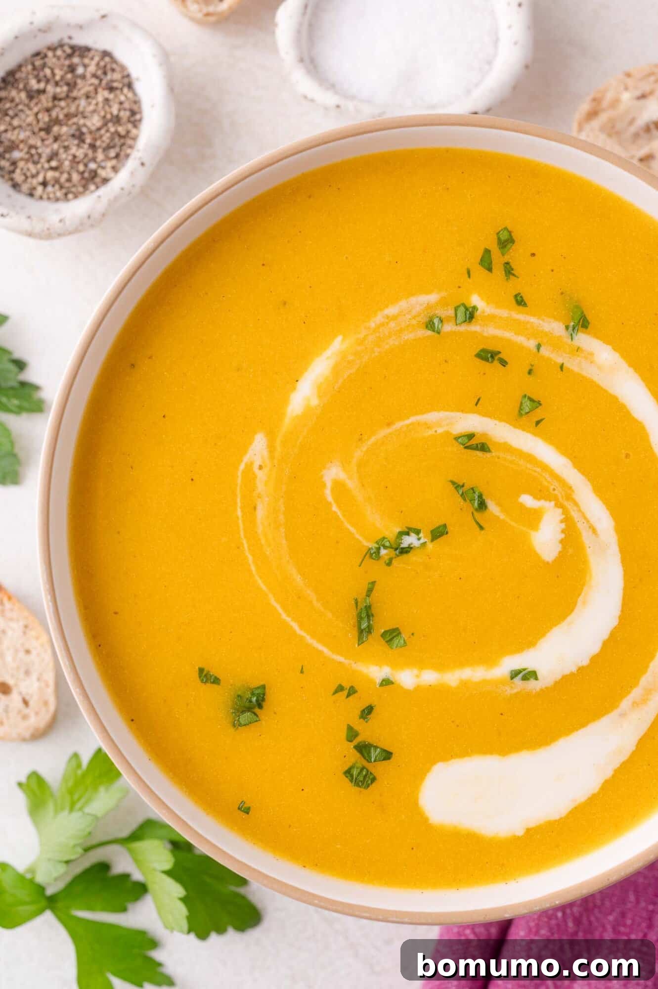 A bowl of creamy kabocha squash soup garnished with a swirl of cream and chopped herbs, surrounded by slices of bread, parsley leaves, and small dishes of salt and pepper.