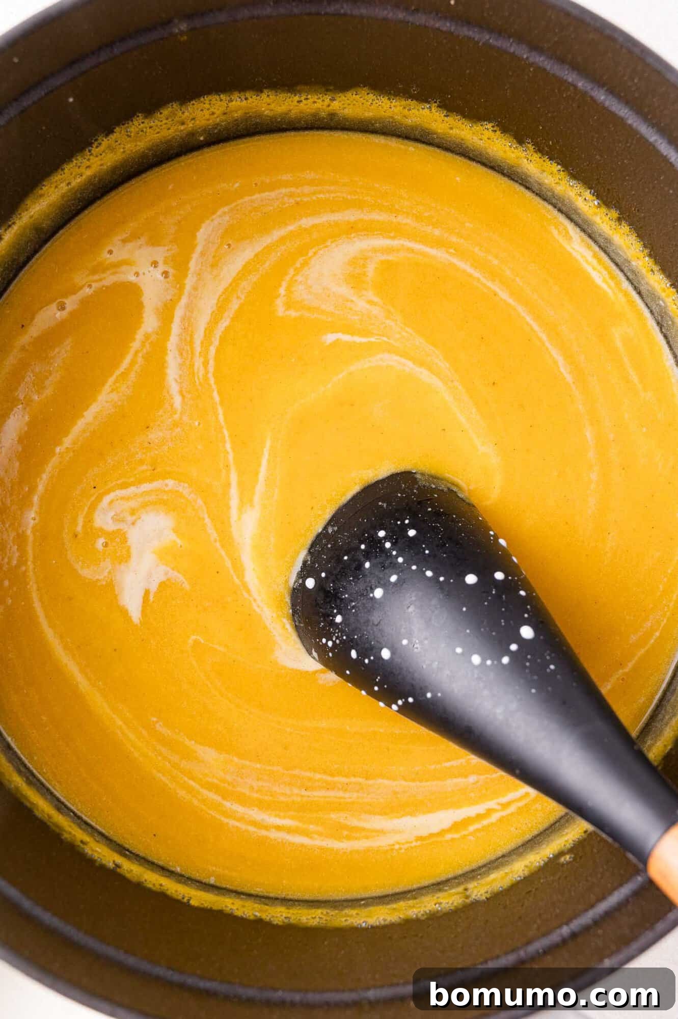 A black spoon stirs a pot of creamy squash soup, showing swirls of white liquid on the surface.