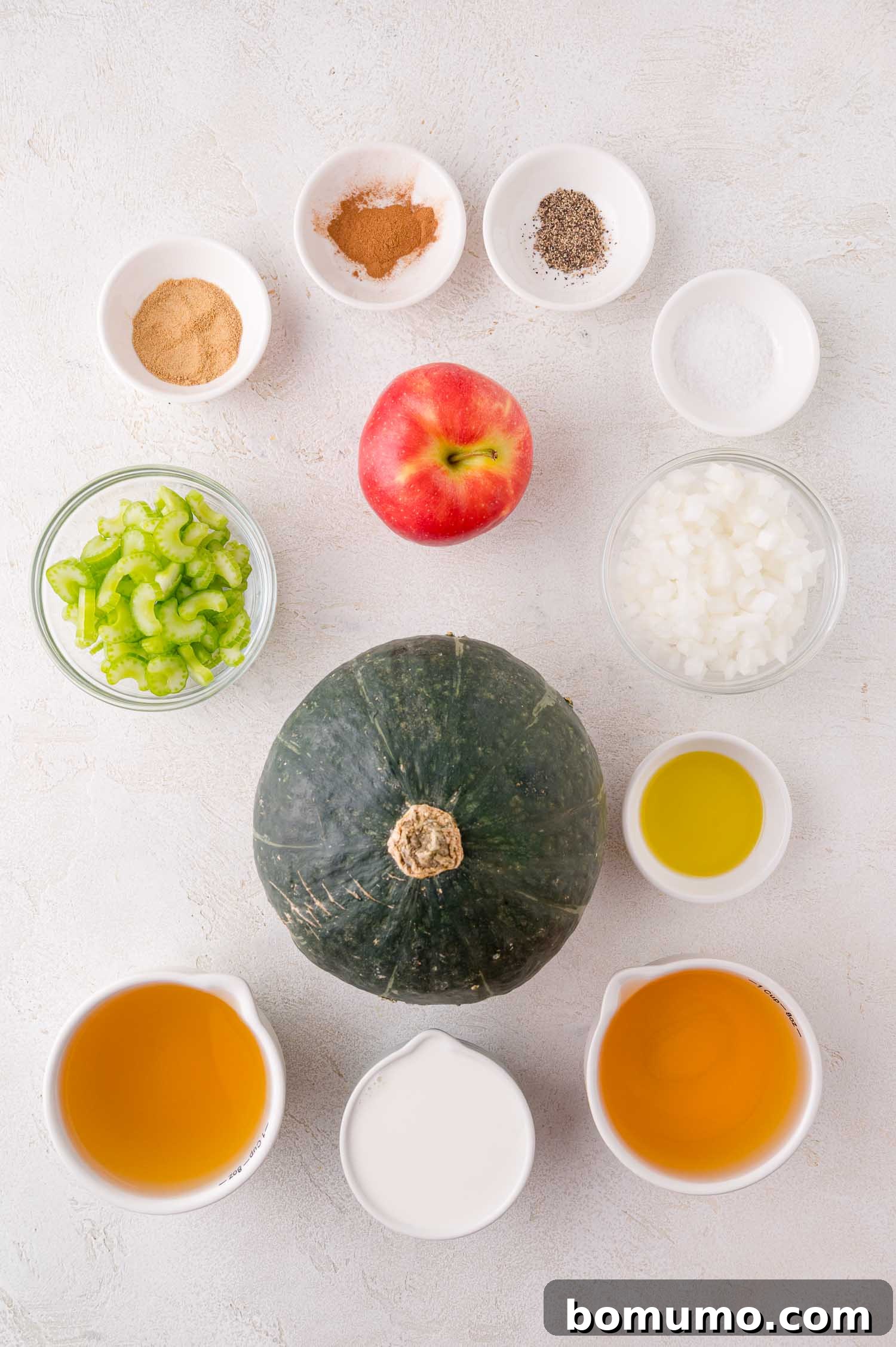 An assortment of ingredients on a white surface, including a whole kabocha squash, apple, celery, onion, spices, broth, olive oil, and cream arranged in separate bowls and cups.