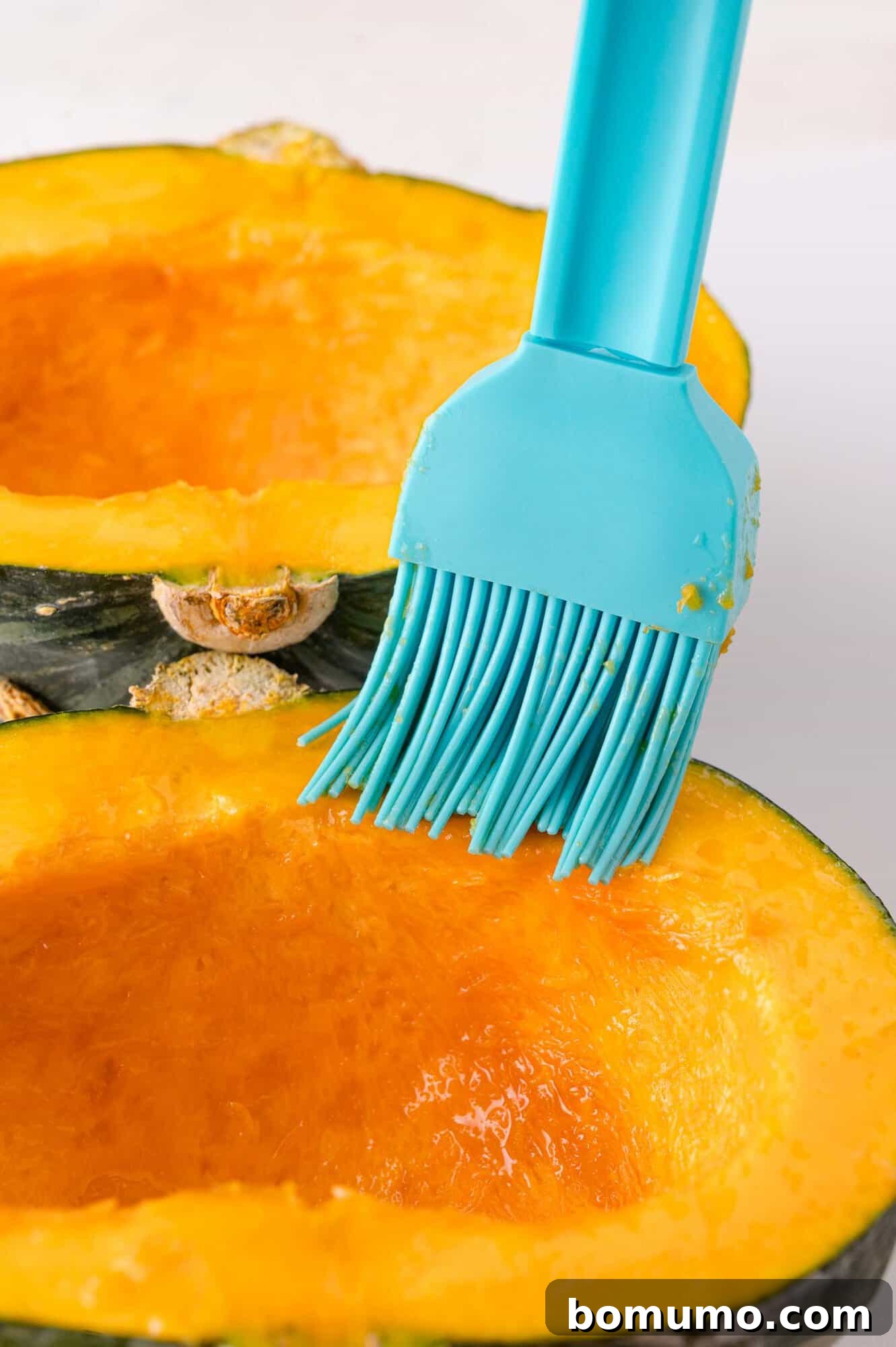 A blue silicone brush applies oil to the cut surface of a halved kabocha squash.