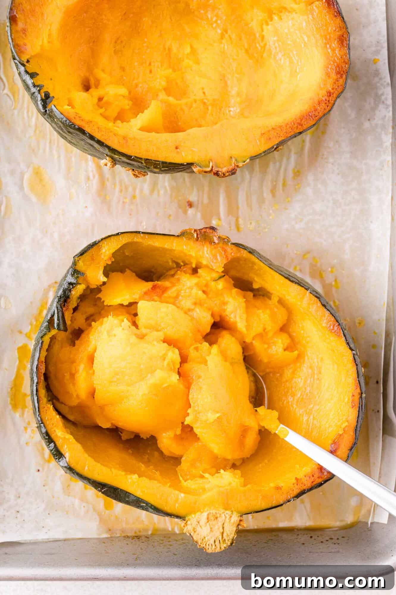 Two halves of a baked kabocha squash on a baking sheet; one half has the flesh scooped out with a spoon.