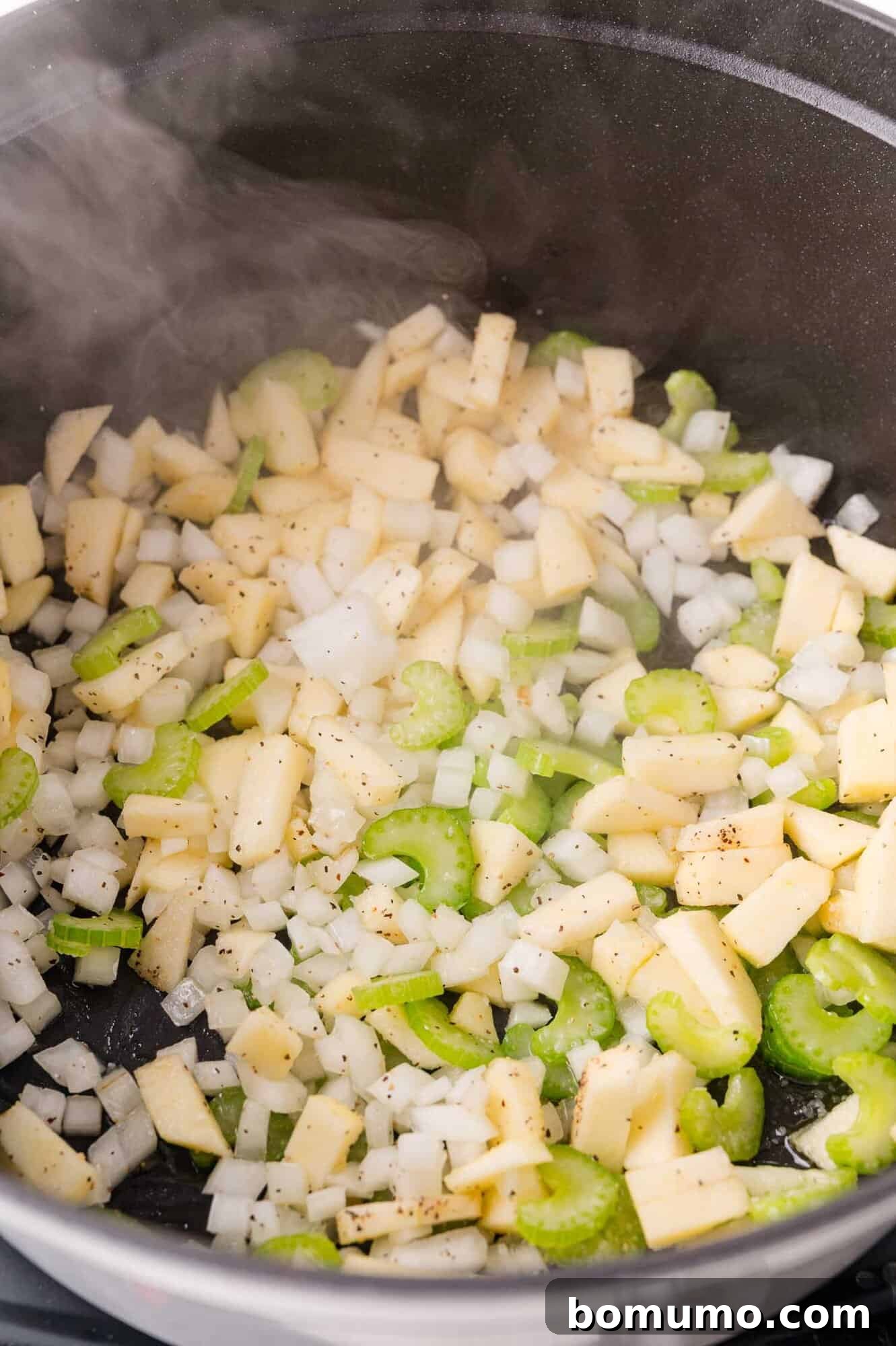 Diced onions, celery, and apples sautéing in a pan with visible steam rising.
