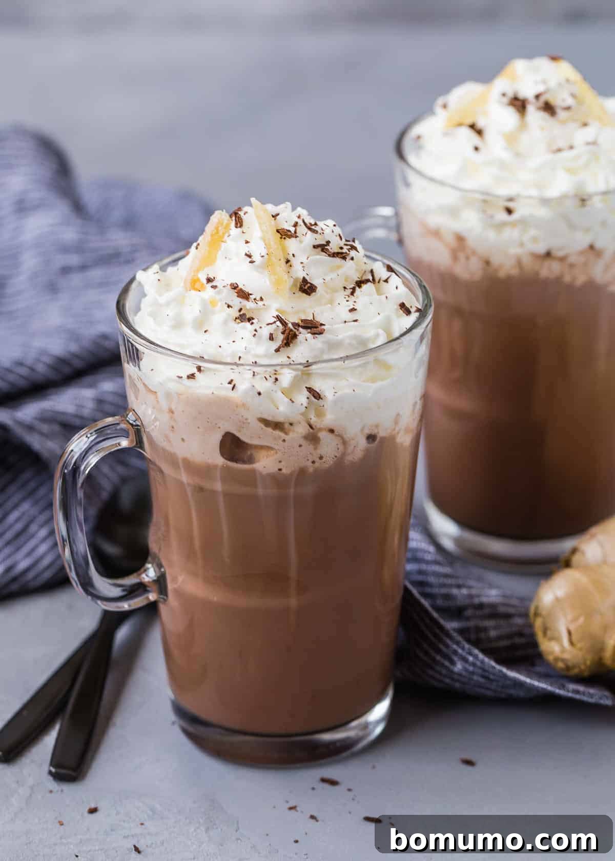 A rustic display of hot cocoa in mugs, alongside black spoons and a fresh knob of ginger, evoking warmth and comfort.