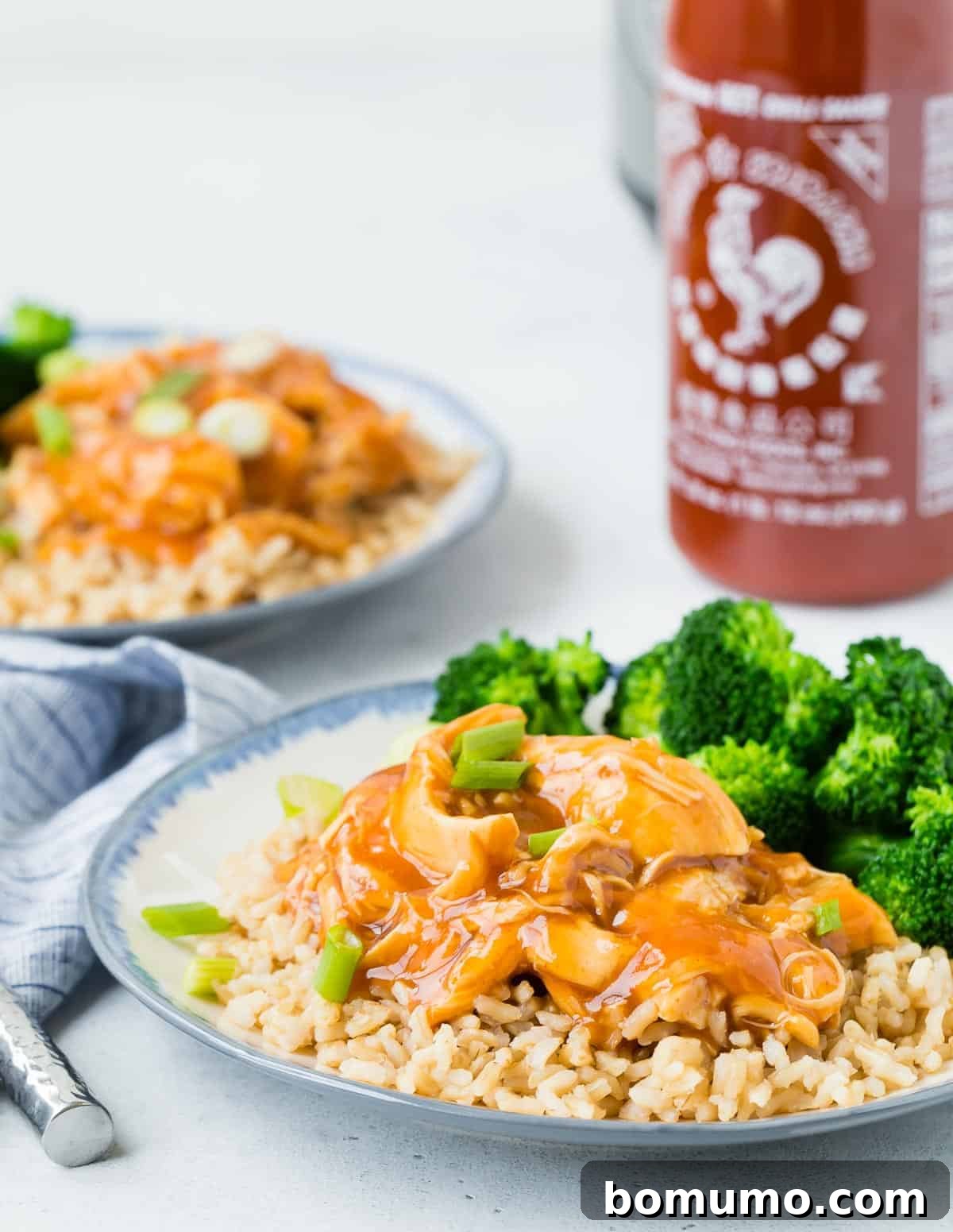 Cooked chicken, rice, and broccoli, with a bottle of sriracha in the background.