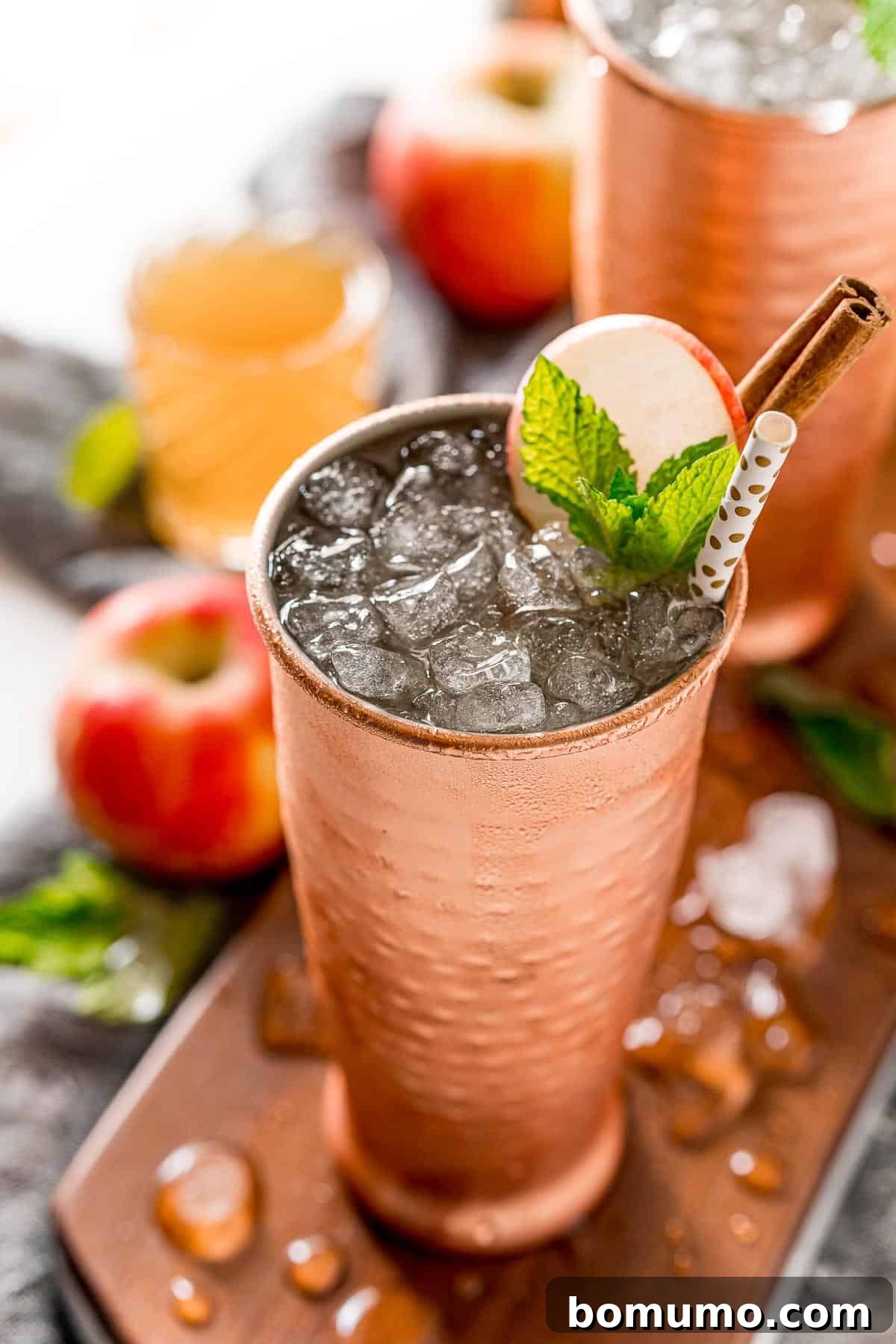 A close-up view of an Apple Cider Moscow Mule filled with nugget ice, elegantly garnished with a fresh apple slice, vibrant mint leaves, and a rustic cinnamon stick.
