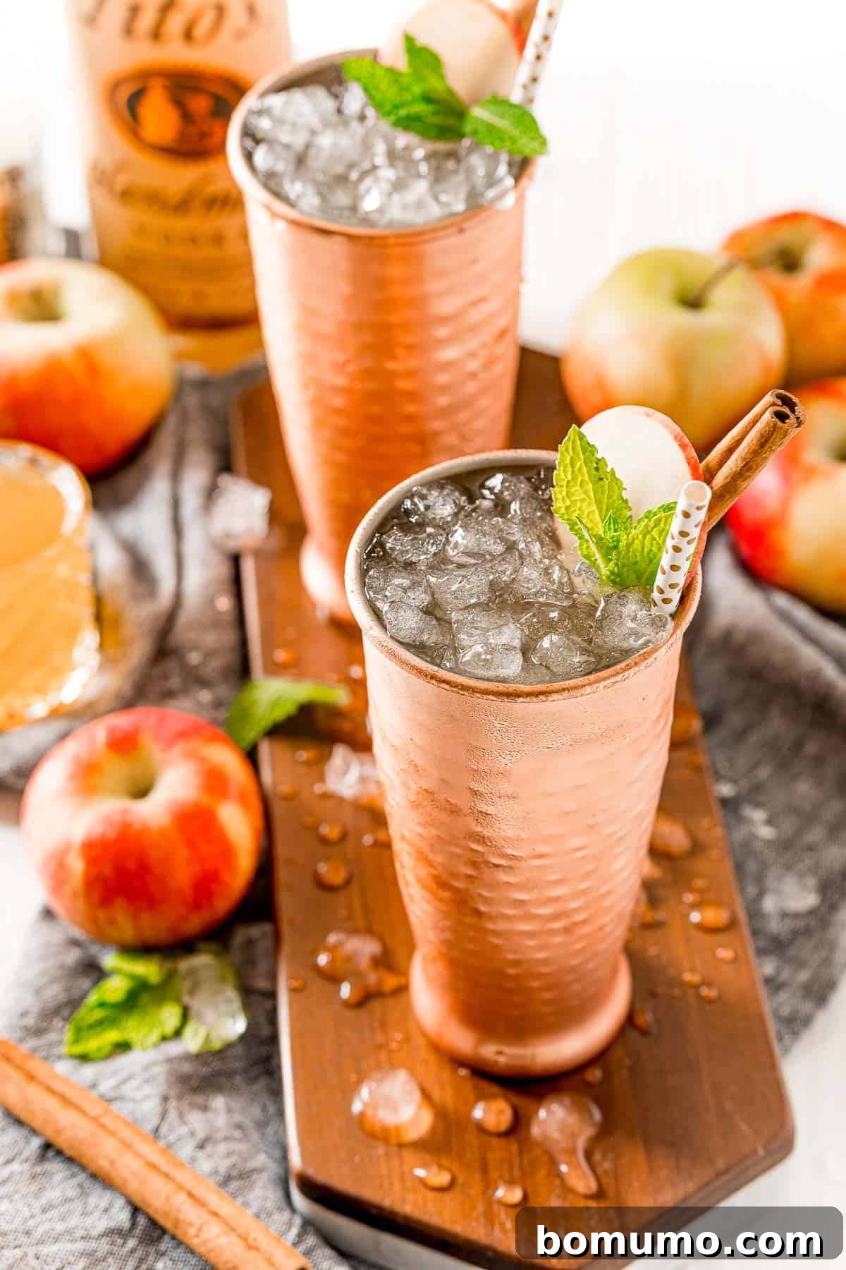 An Apple Cider Moscow Mule, brimming with nugget ice, is artfully garnished with a crisp apple slice, aromatic mint leaves, and a fragrant cinnamon stick, perfect for a festive autumn celebration.