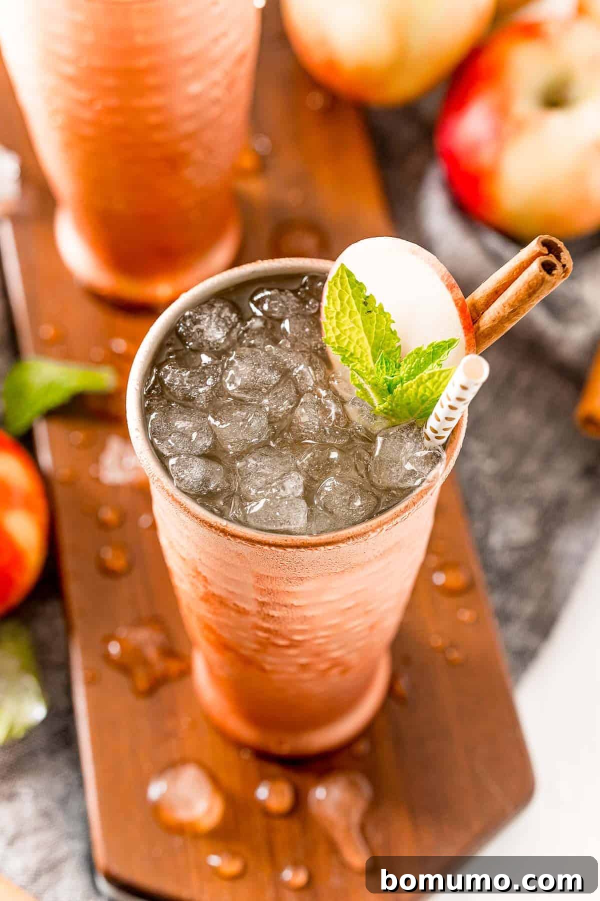 A festive Apple Cider Moscow Mule, presented with nugget ice and adorned with a vibrant apple slice, fresh mint leaves, and a fragrant cinnamon stick, captures the essence of autumn in a single glass.