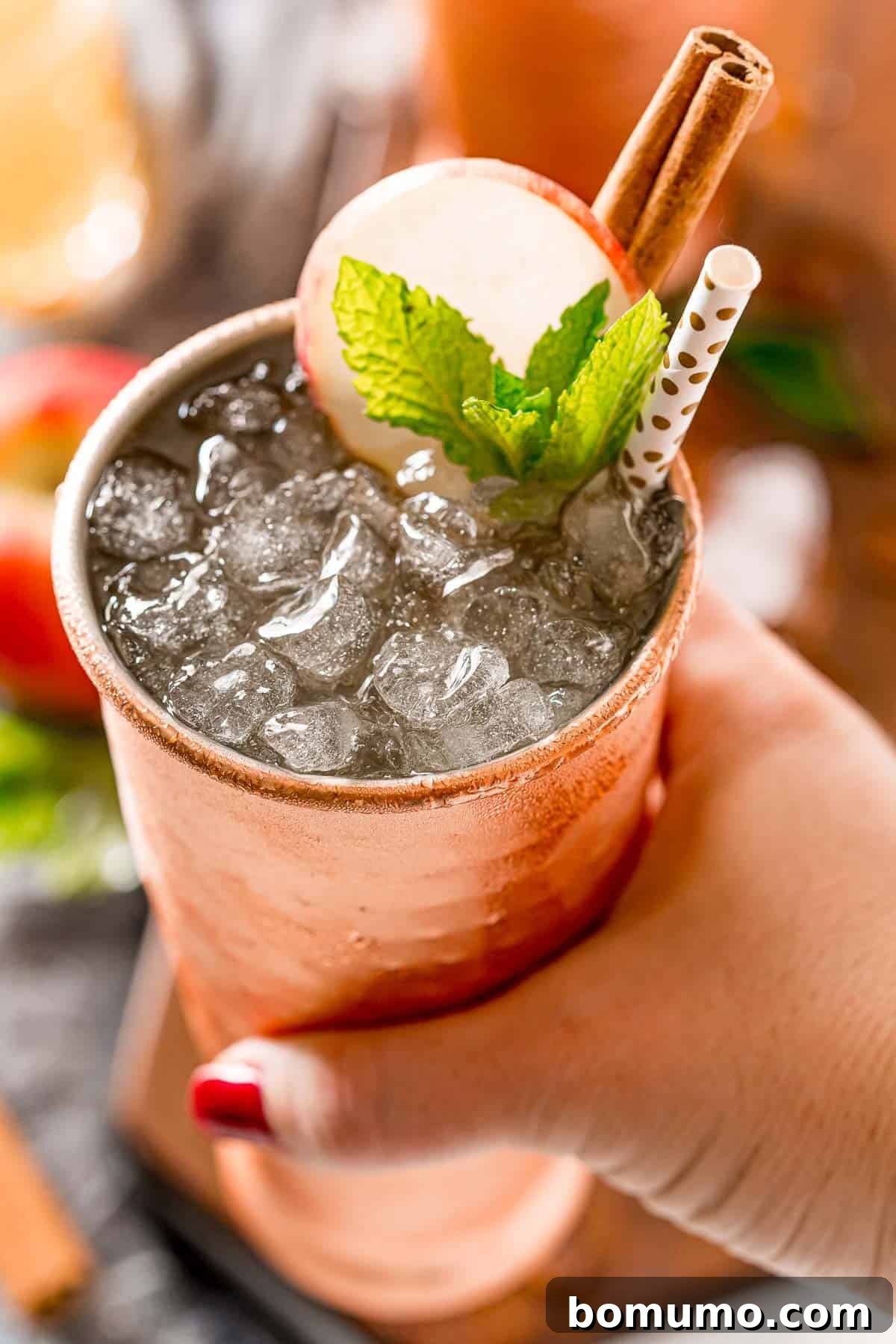 A woman's hand gracefully holding an Apple Cider Moscow Mule in a copper mug, highlighting the festive drink in an inviting setting.
