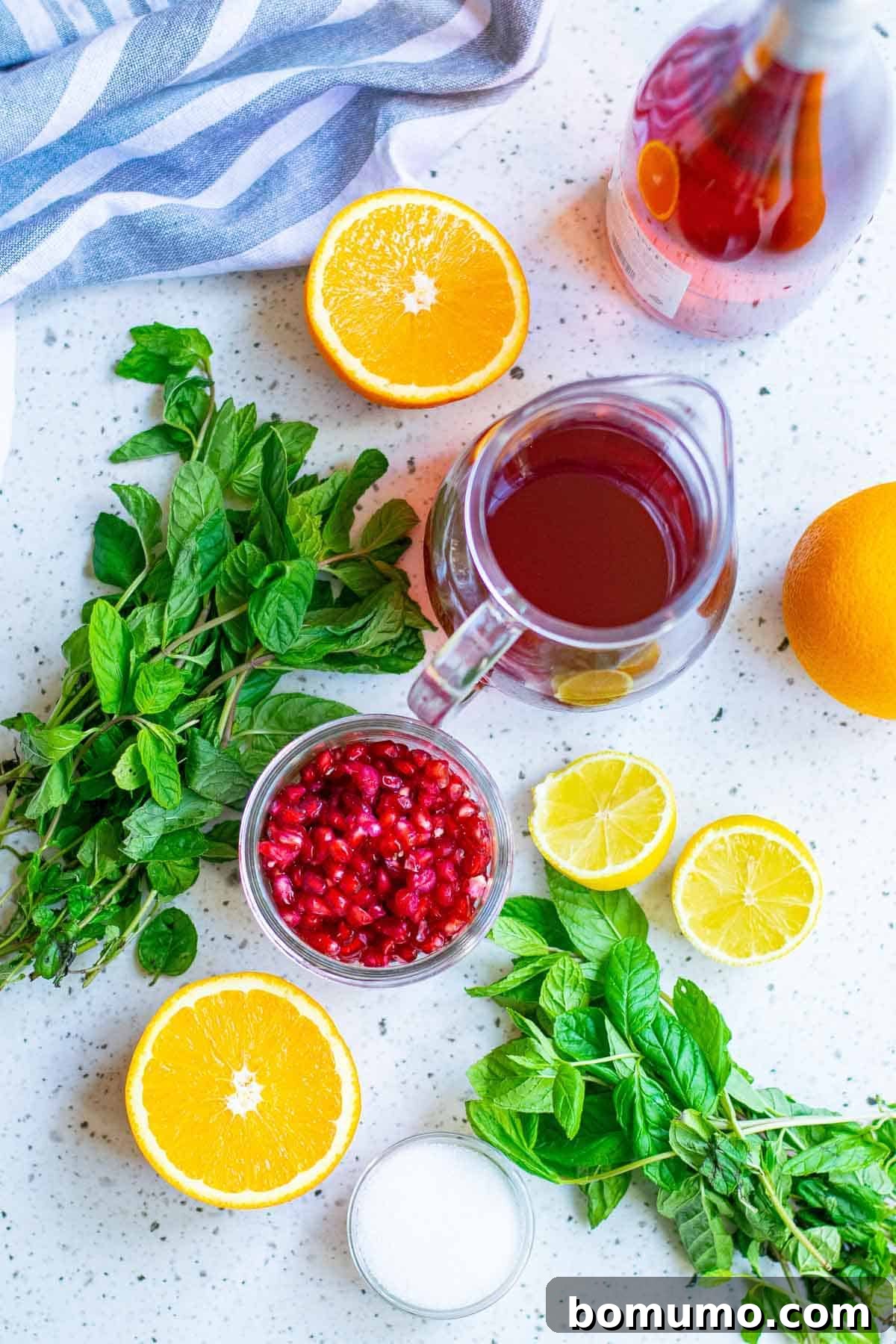 Overhead view of all fresh ingredients laid out for sparkling pomegranate punch: pomegranate juice, sugar, citrus slices, pomegranate arils, sparkling wine, and fresh mint.