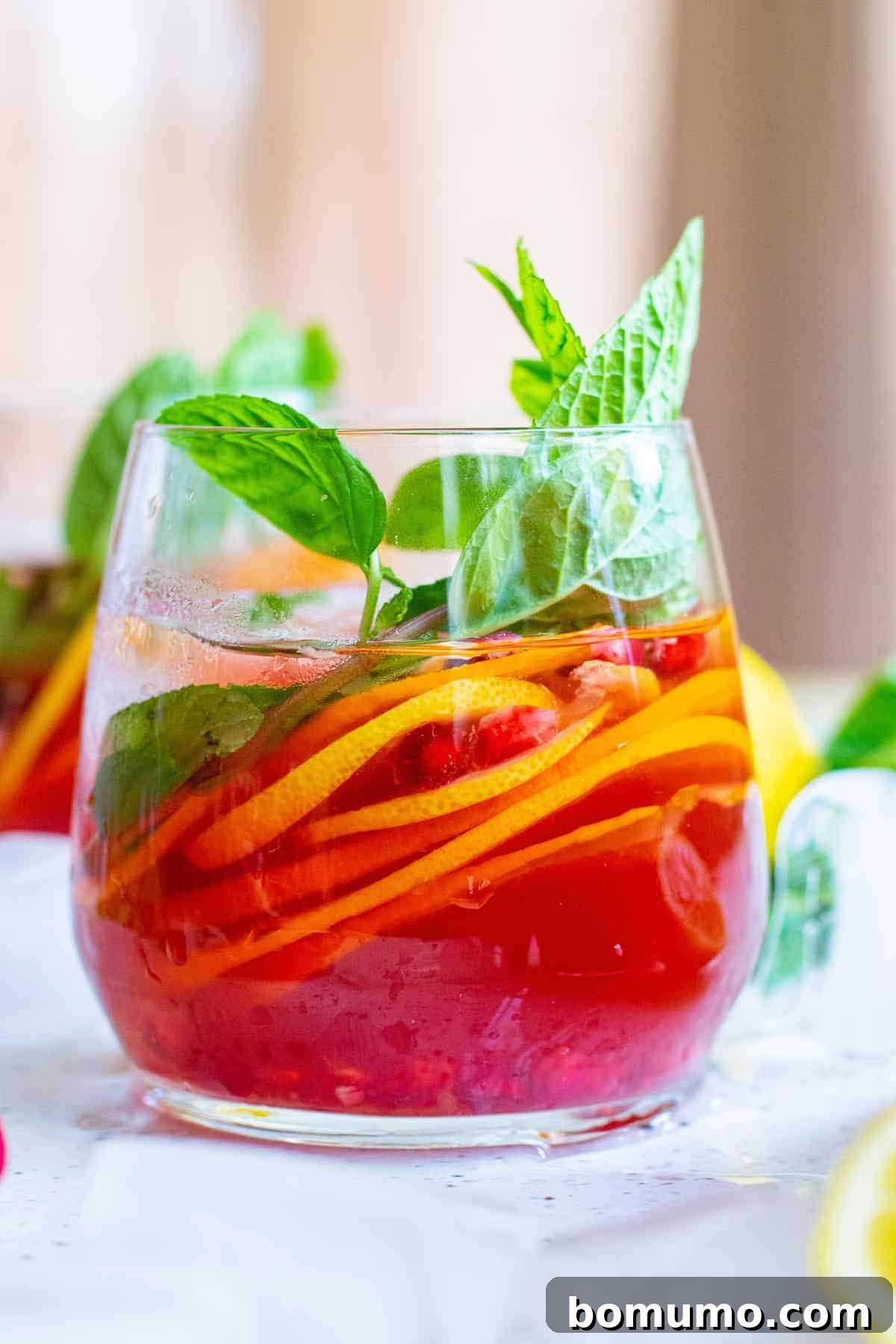 Close-up view of a stemless glass filled with vibrant ruby-red sparkling pomegranate punch, garnished with glistening arils, bright citrus slices, and fresh mint leaves.
