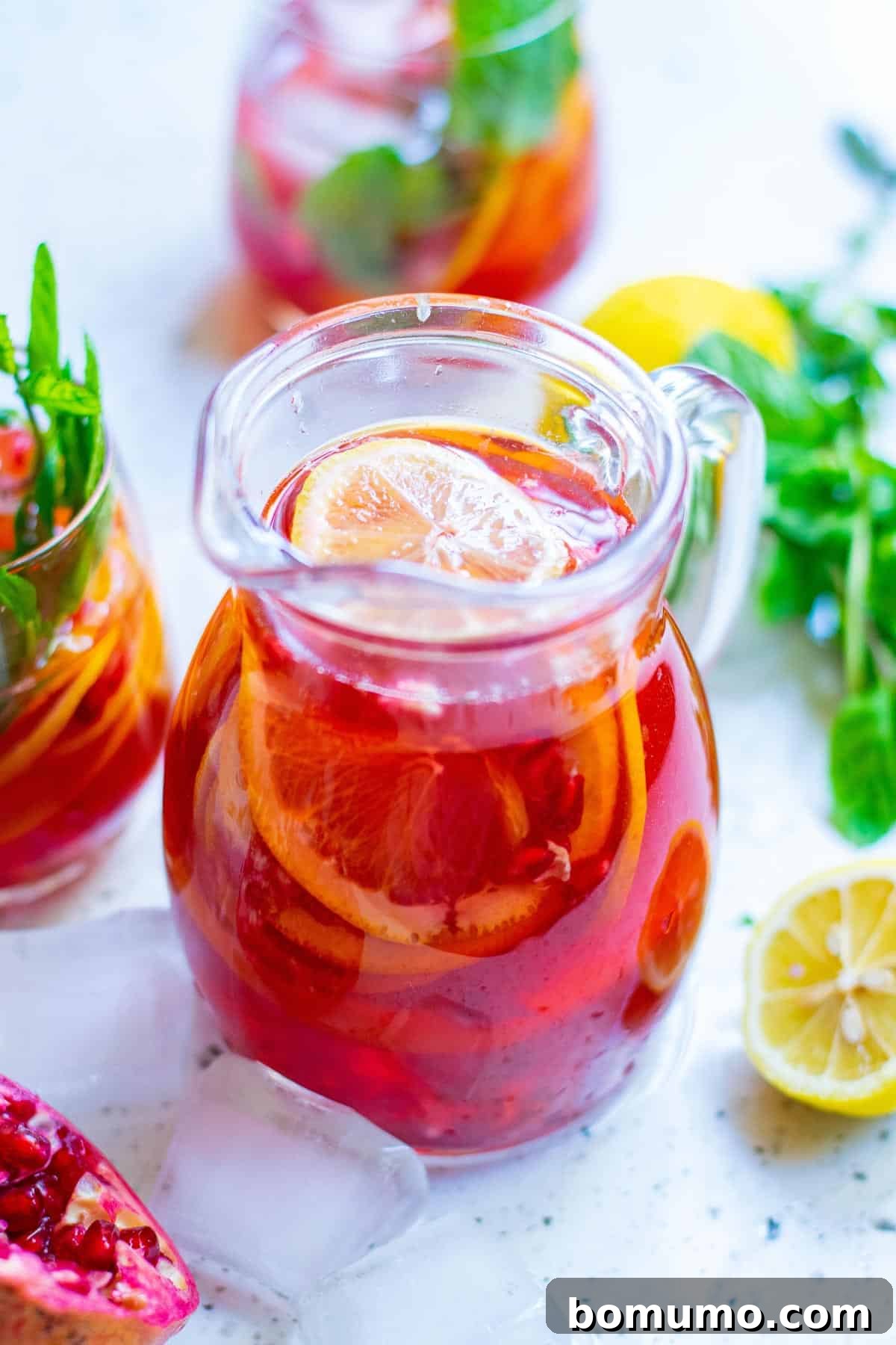 A large glass pitcher of bright red punch, beautifully adorned with colorful fresh garnishes including citrus slices, pomegranate arils, and mint leaves, ready for pouring.