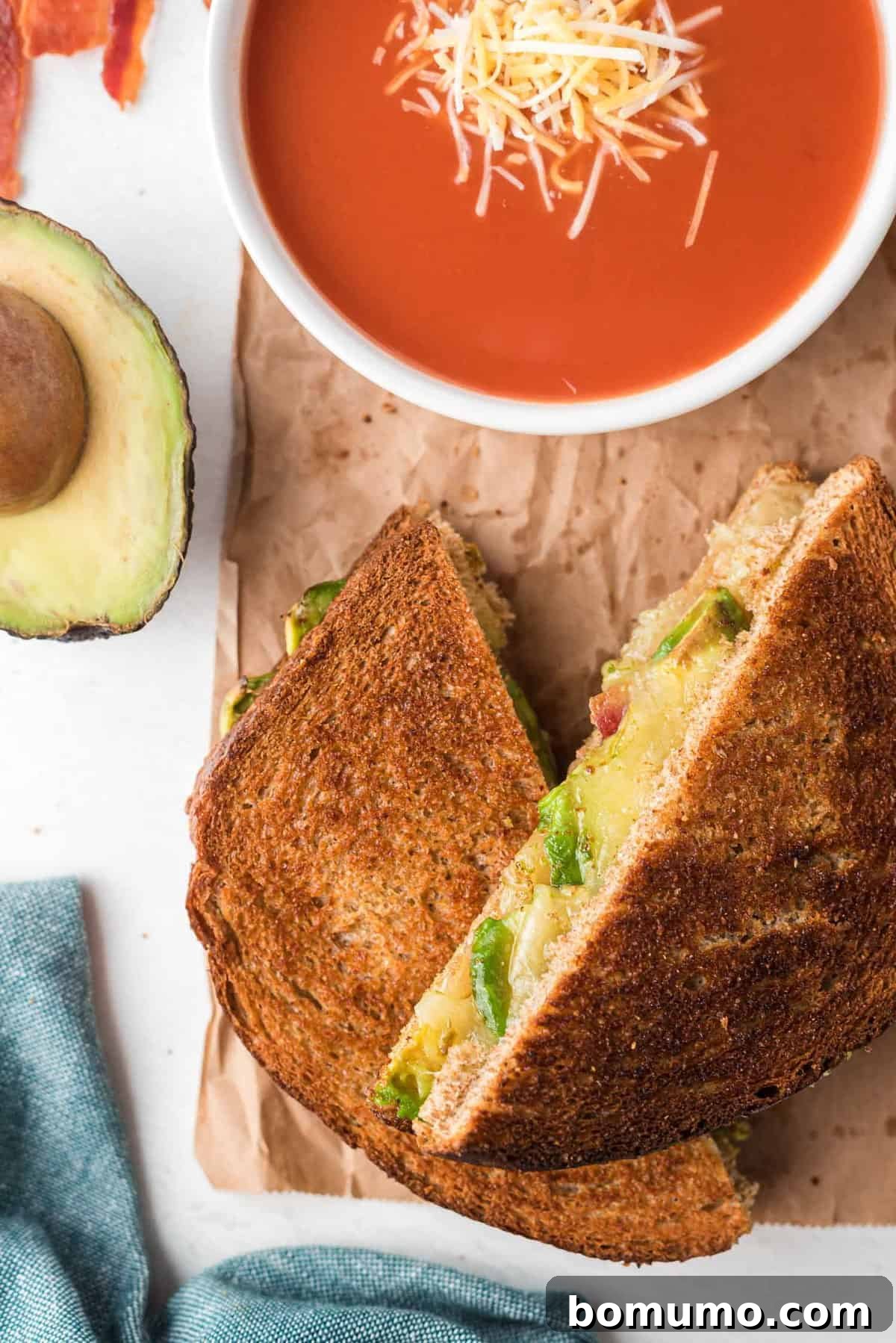 Sandwich halves that halve been toasted, filled with avocado, cheese, and bacon, served with tomato soup.
