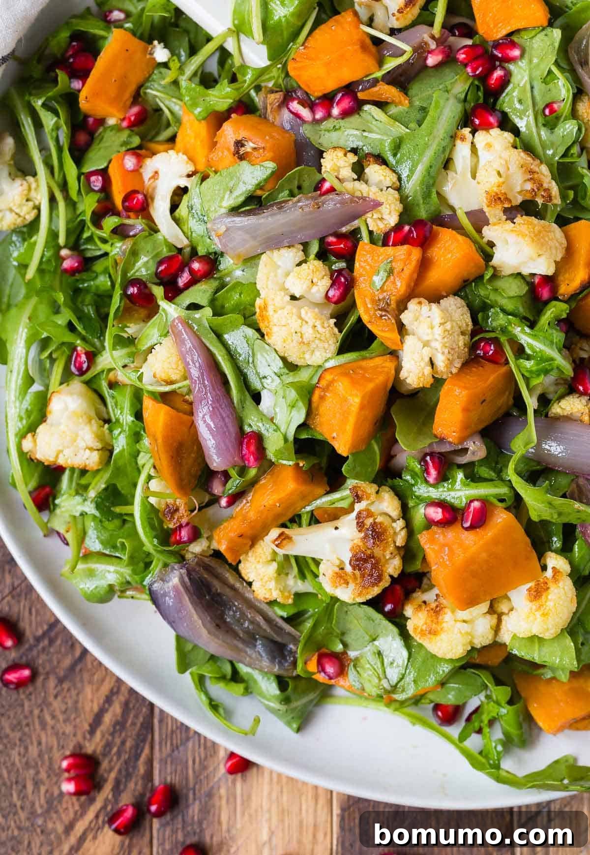 Fresh arugula arranged on a large white platter, forming a base for a generous topping of roasted cauliflower and roasted sweet potatoes, awaiting final assembly.