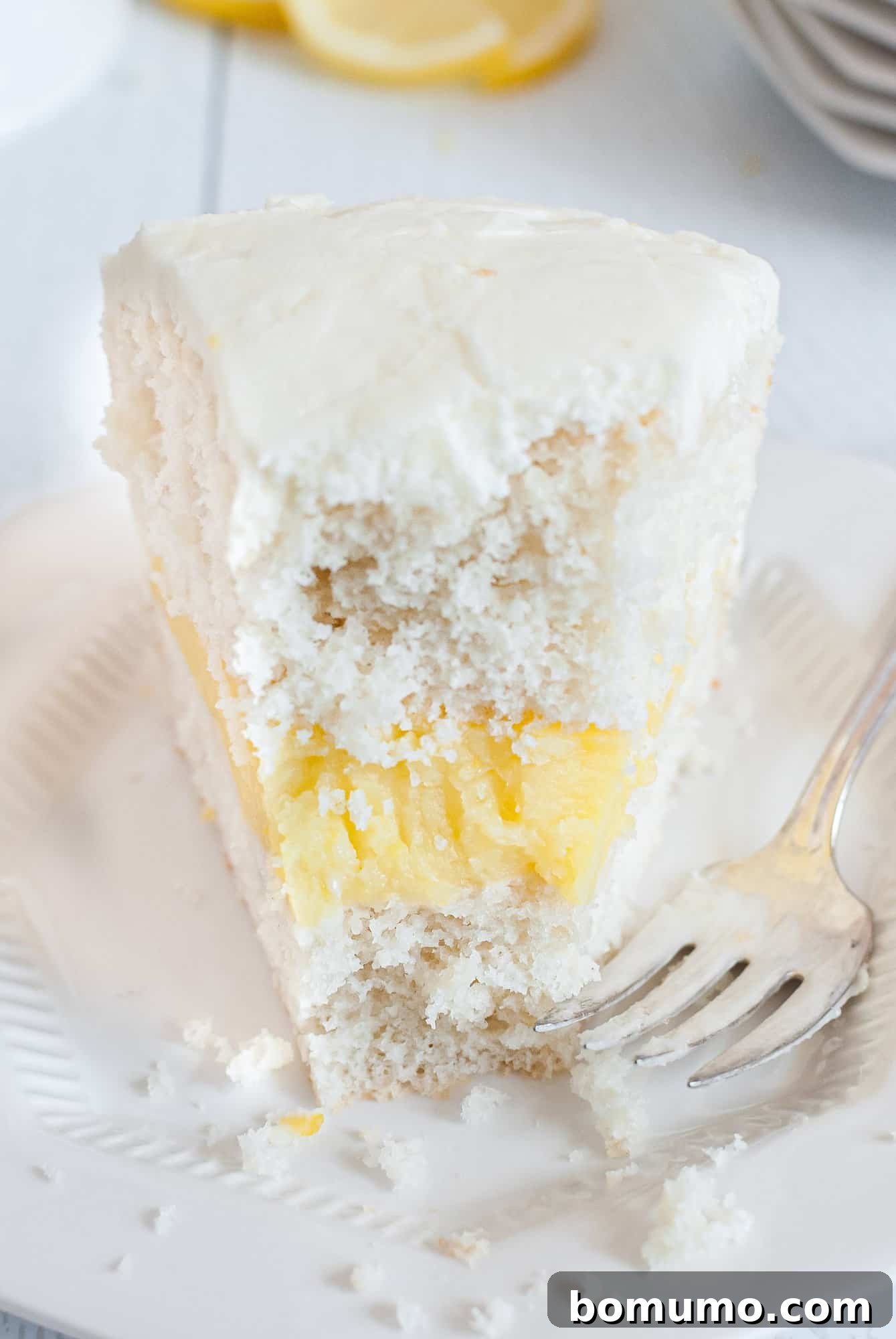 A close-up of a slice of lemon cake, revealing its vibrant lemon filling and creamy frosting, with a small bite removed.