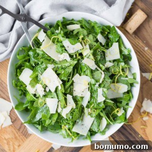 Overhead view of a white bowl with an arugula salad featuring Parmesan and lemon, set against a warm wooden background.